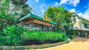 Un restaurante llamado 'Amazon' con techo verde, rodeado de abundante vegetación y un pequeño estanque con peces.