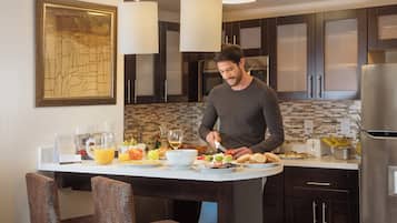 A modern kitchen with dark cabinetry, a marble countertop, and a breakfast setup including fruits, bread, and a wine glass.