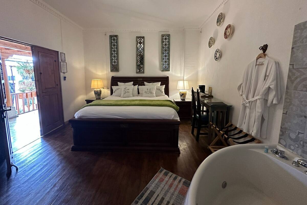 Une chambre bien éclairée avec un grand lit, un plancher en bois, une table de chevet avec une lampe, une chaise, un peignoir accroché au mur et une baignoire.