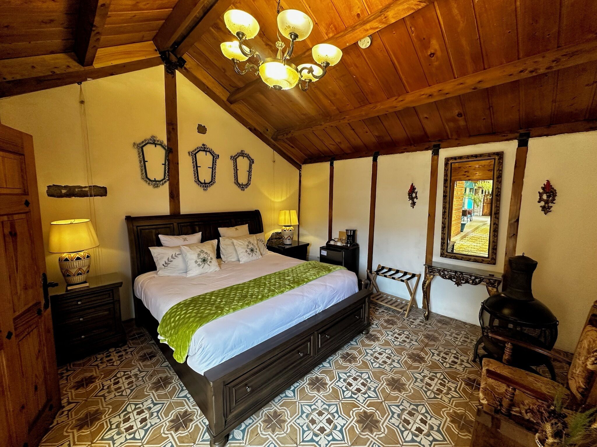 A bedroom with a large bed, wooden ceiling beams, patterned floor tiles, and a chandelier.