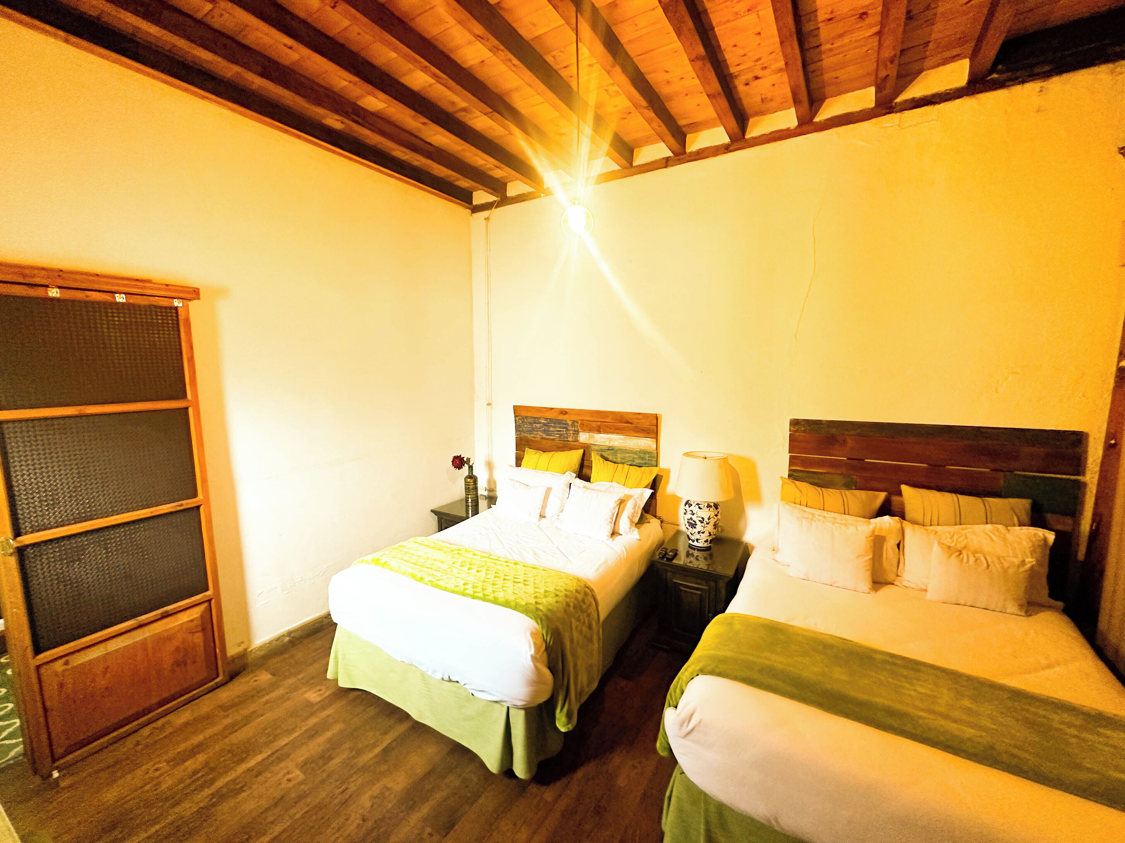 Una habitación con dos camas, vigas de madera en el techo, una mesita con una lámpara y una ventana con persianas.