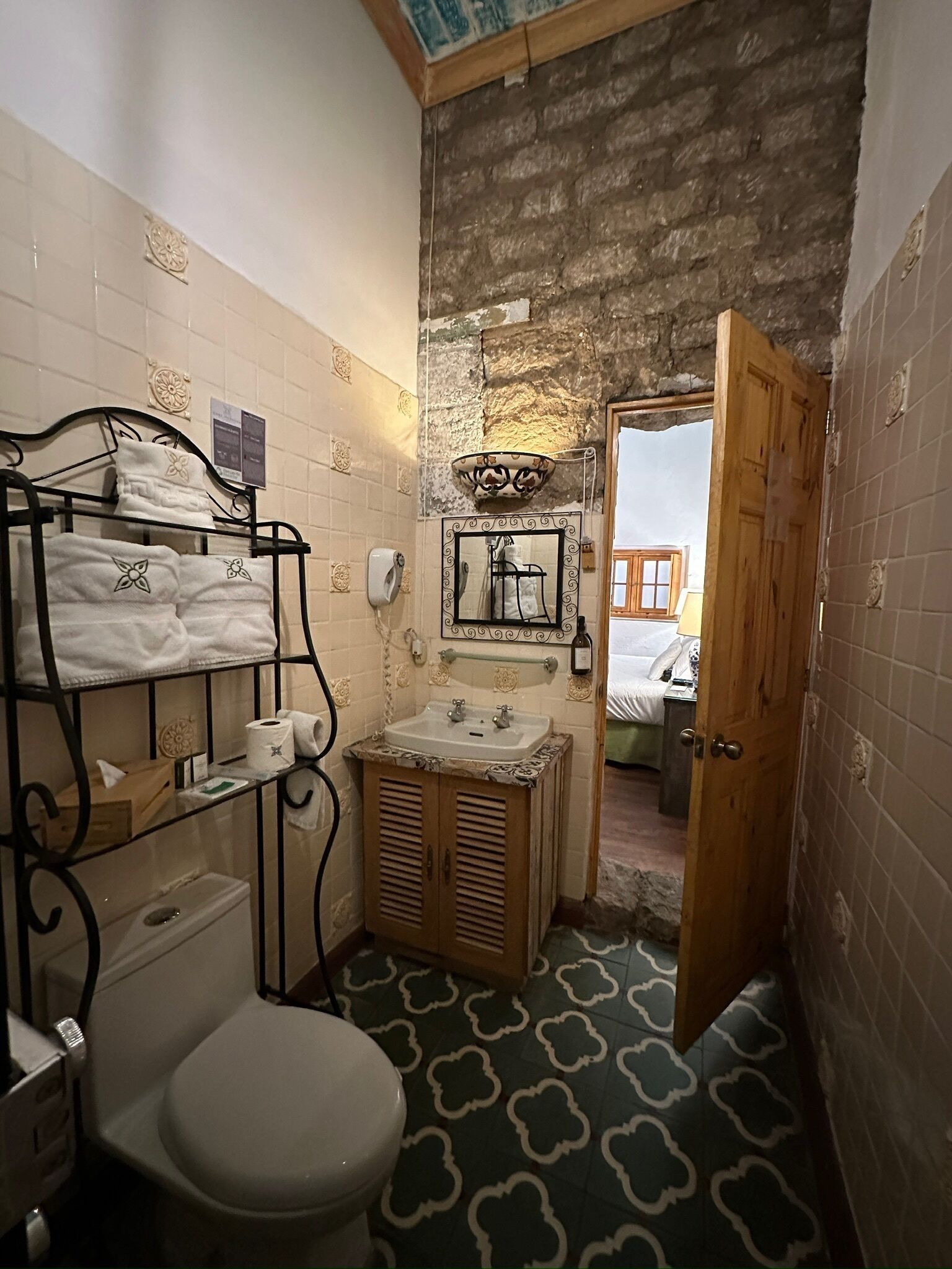 Un baño pequeño con inodoro, lavamanos y toallero. Una puerta de madera da paso a otra habitación.