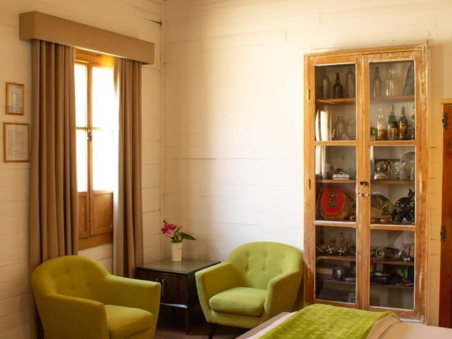 Una habitación con cama, dos sillones, un aparador de madera con puertas de cristal, una ventana con cortinas y una lámpara colgante.