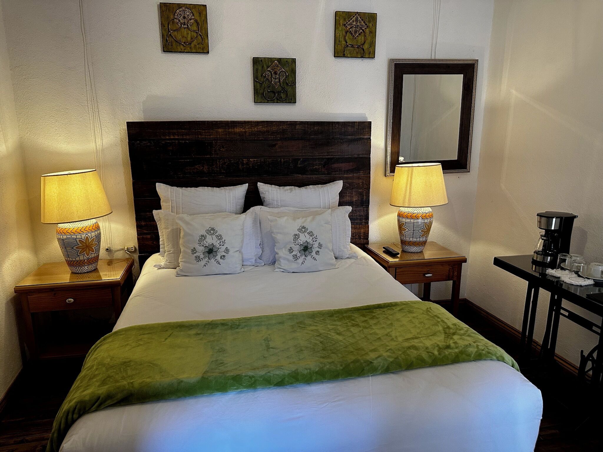 A neatly made bed with a green throw blanket, two bedside lamps, and a coffee maker on a small table beside the bed.
