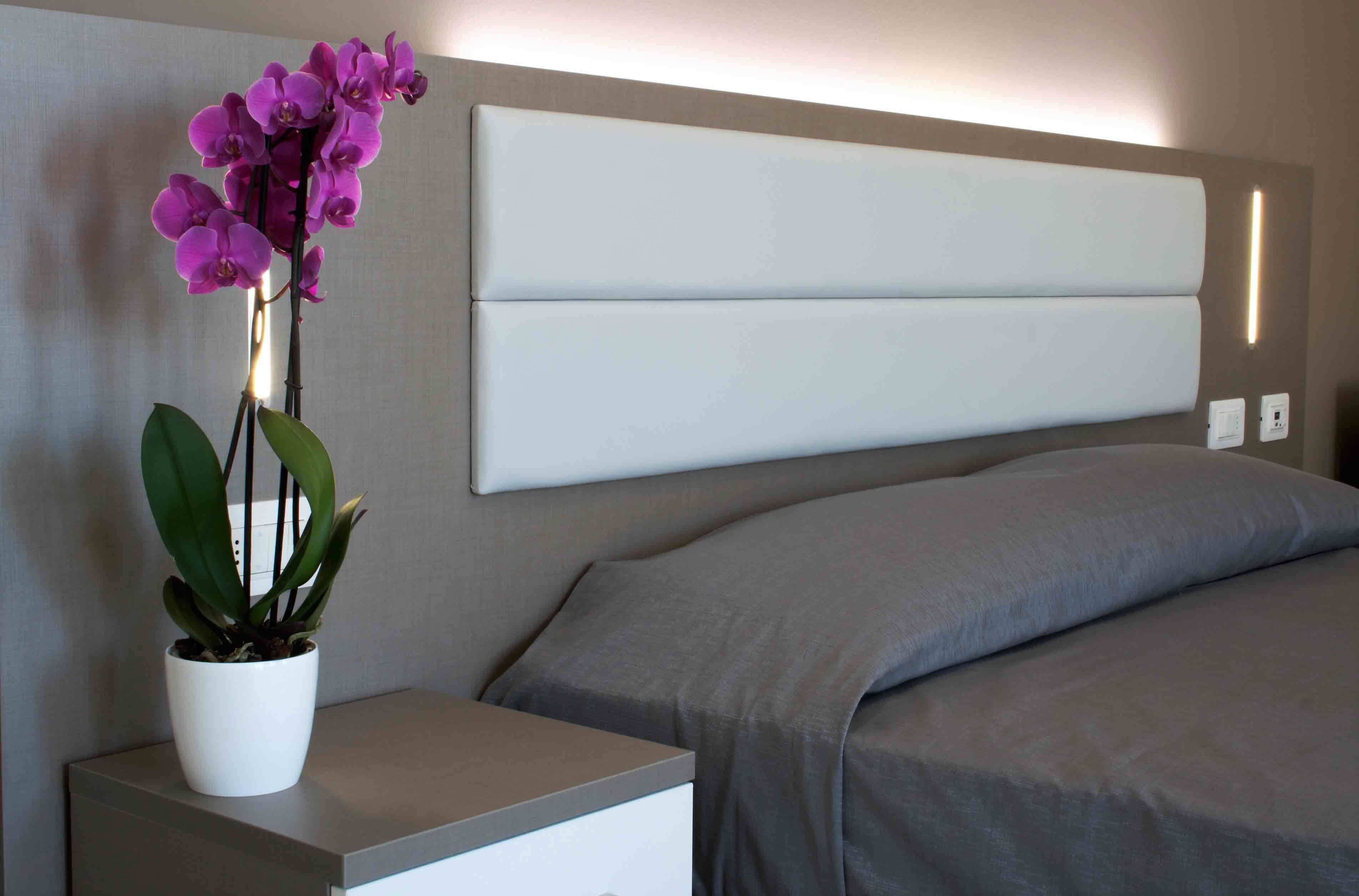 A modern hotel room with a bed, a nightstand with a potted orchid, and a wall-mounted light fixture.