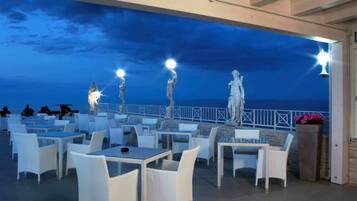 An outdoor seating area with tables and chairs, a view of the sea, and decorative statues.