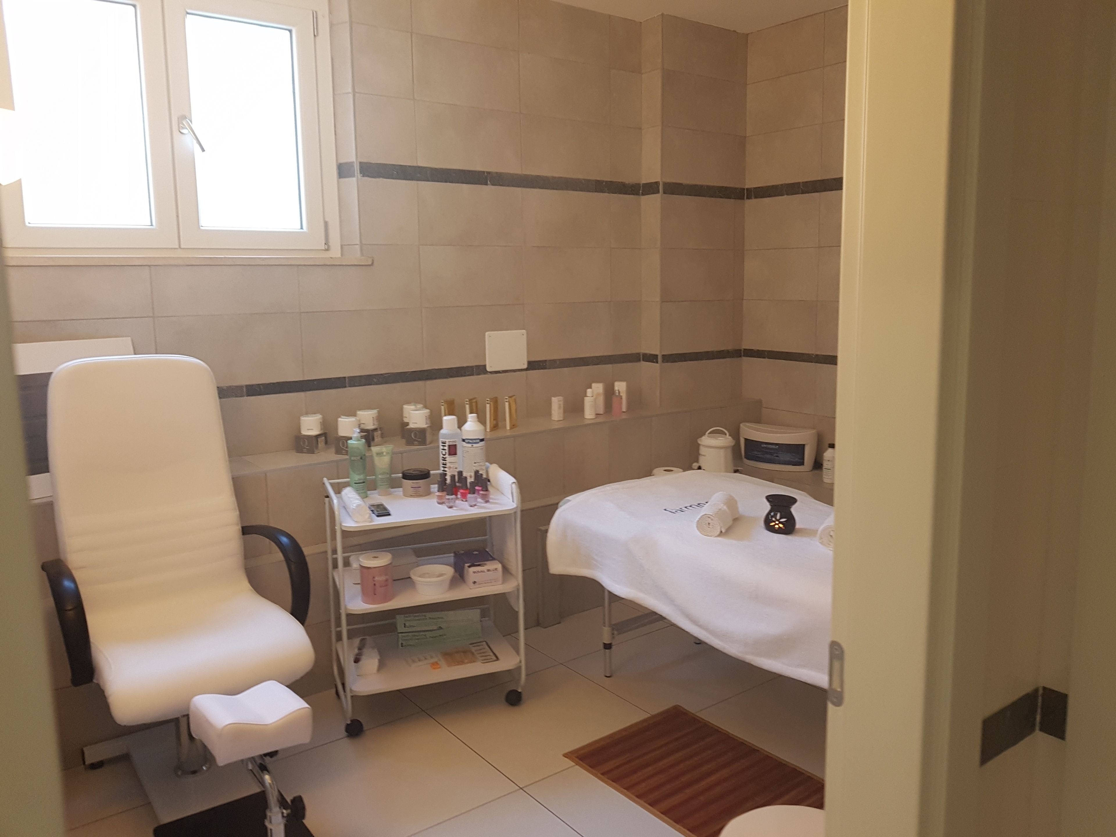 A bathroom with a white chair, a shelf with toiletries, a bed with a white towel, and a window.