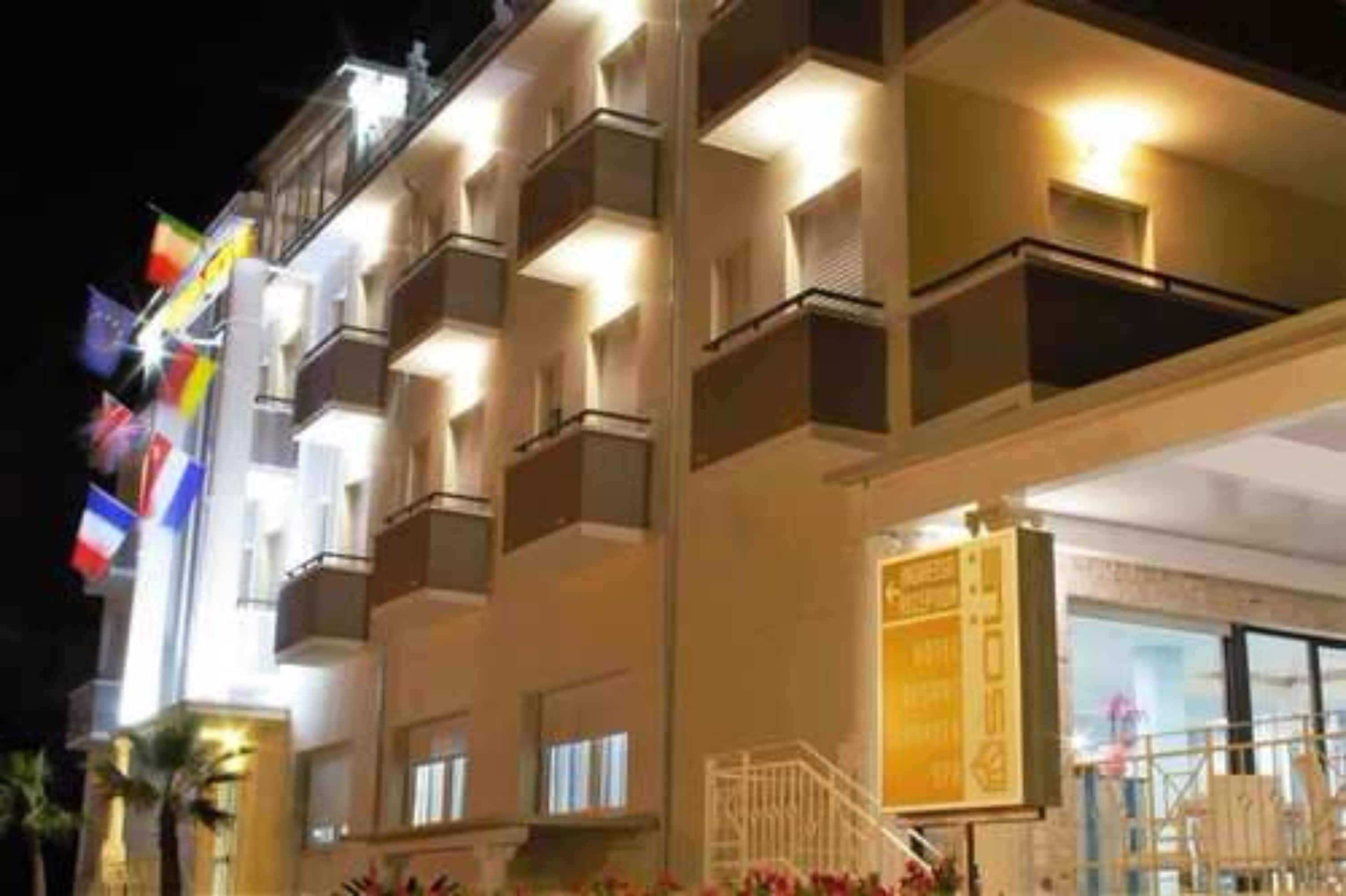 Un hotel de varios pisos con balcones, una entrada bien iluminada y banderas en el exterior.