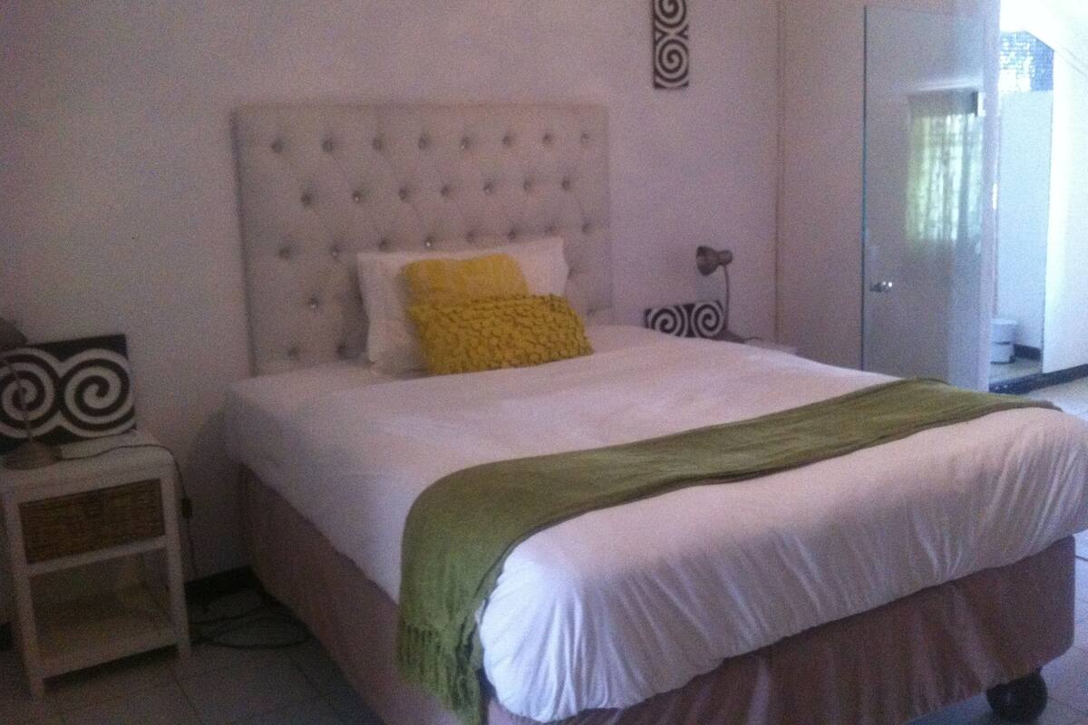 Une chambre avec une tête de lit capitonnée, un lit avec une literie blanche et jaune, un plaid vert, une table de chevet agrémentée d’un objet décoratif et une porte donnant sur une autre pièce.