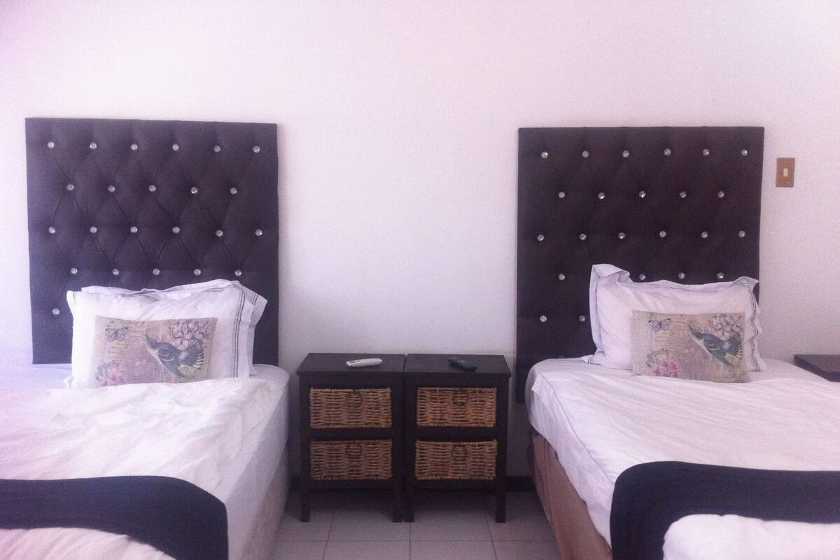 Une chambre d’hôtel avec deux lits, chacun doté d’une tête de lit sombre et de draps blancs. Une petite table avec des paniers se trouve entre les lits.