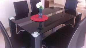 Une table de salle à manger rectangulaire, dotée d’un plateau en verre et de quatre chaises noires. Un petit vase contenant une fleur blanche est placé au centre de la table.
