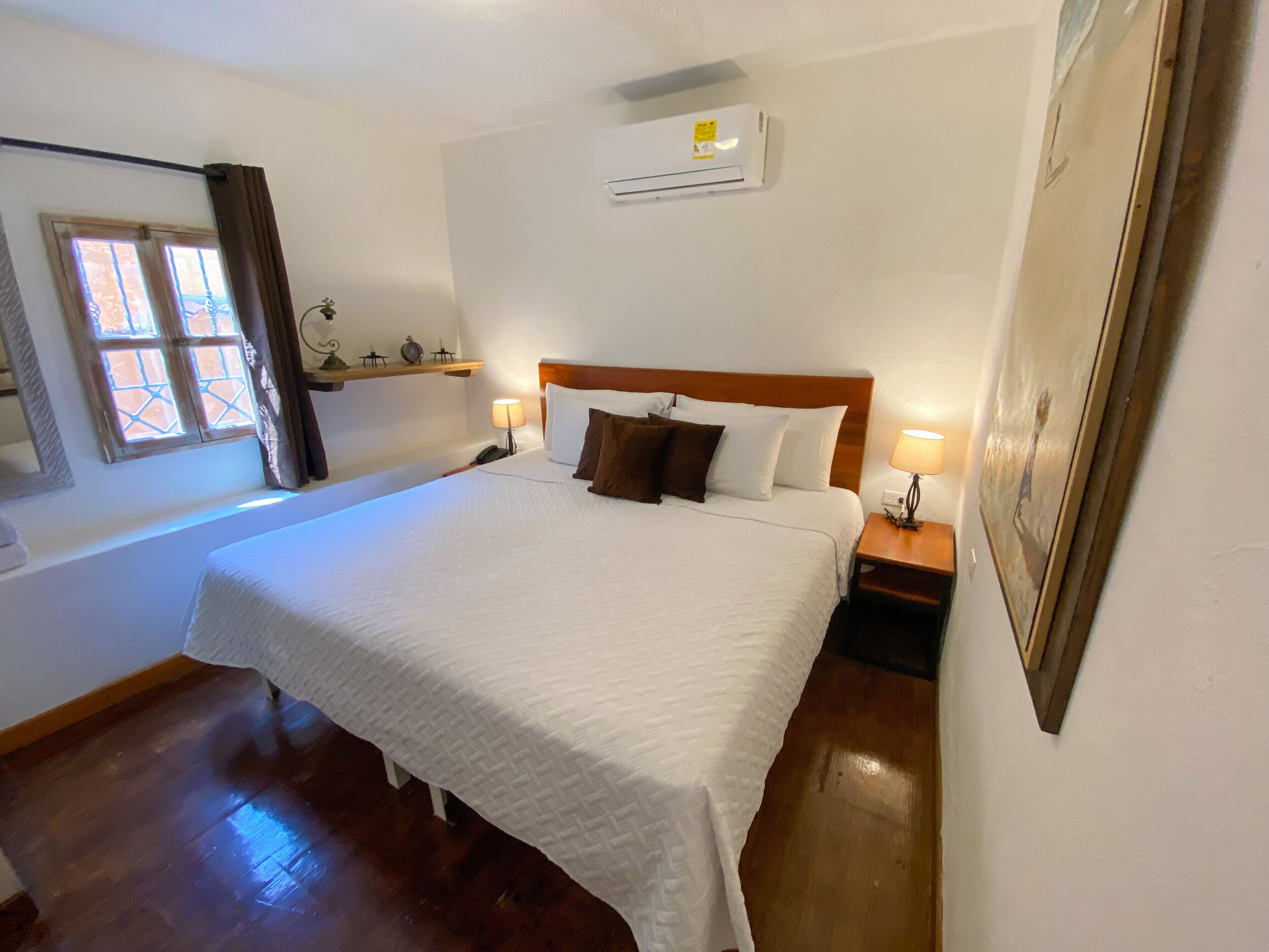 A neatly made bed with white linens and brown pillows, a bedside lamp, and a wall-mounted air conditioner.