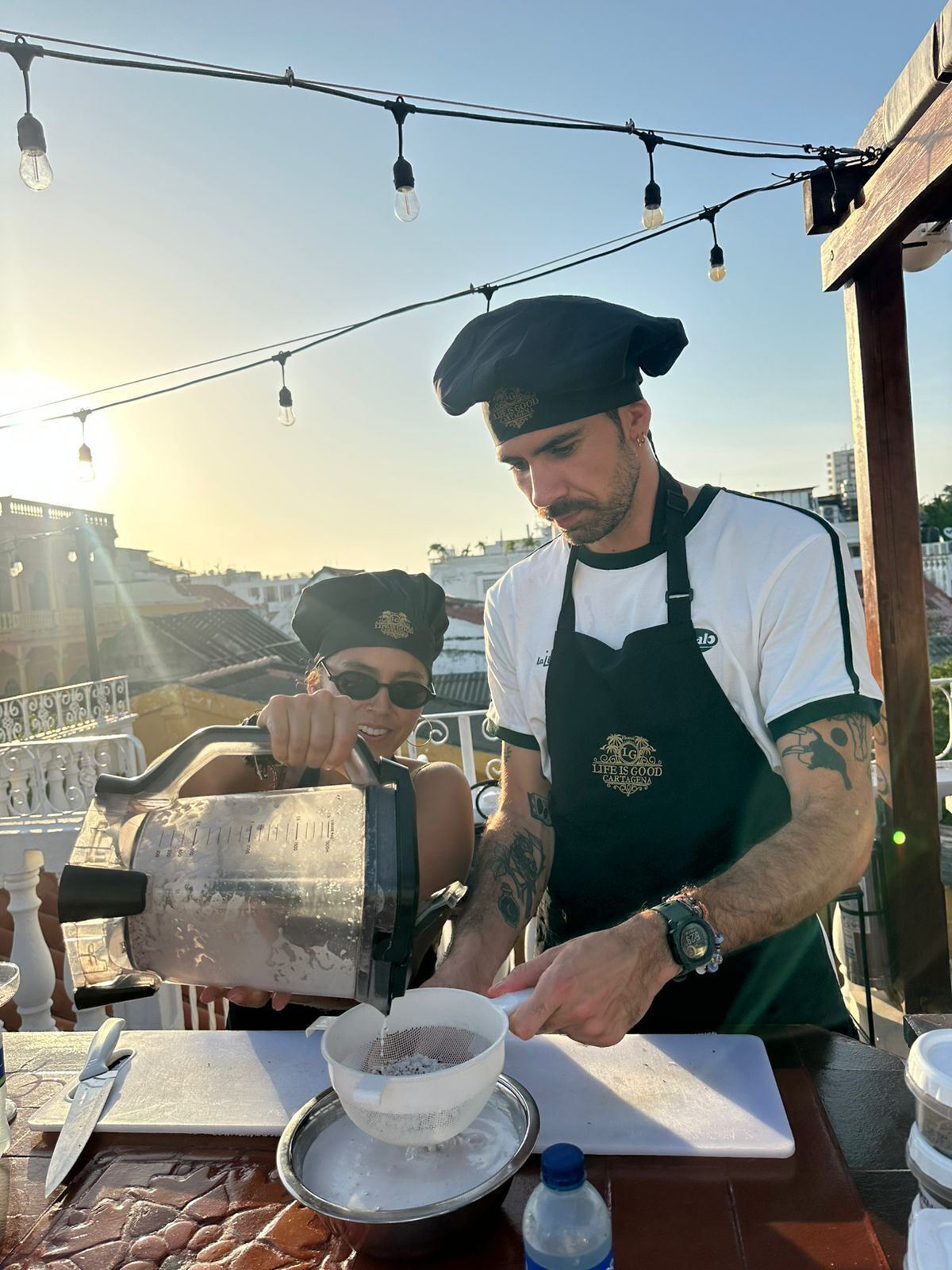 A person preparing coffee with a manual espresso machine, a water bottle, and a cup on a table with a view of buildings in the background.