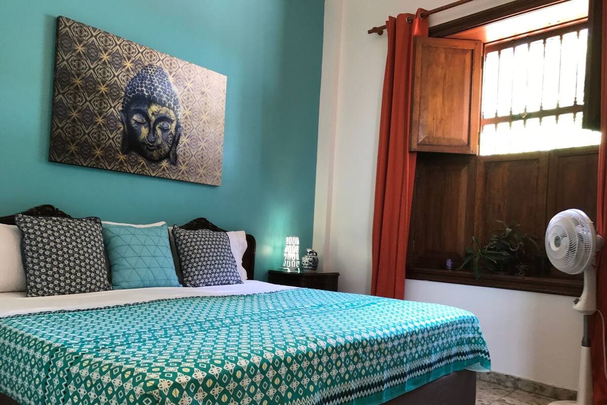 A bedroom with a bed, patterned bedding, a wall-mounted Buddha painting, a bedside table with a lamp, and a window with curtains.