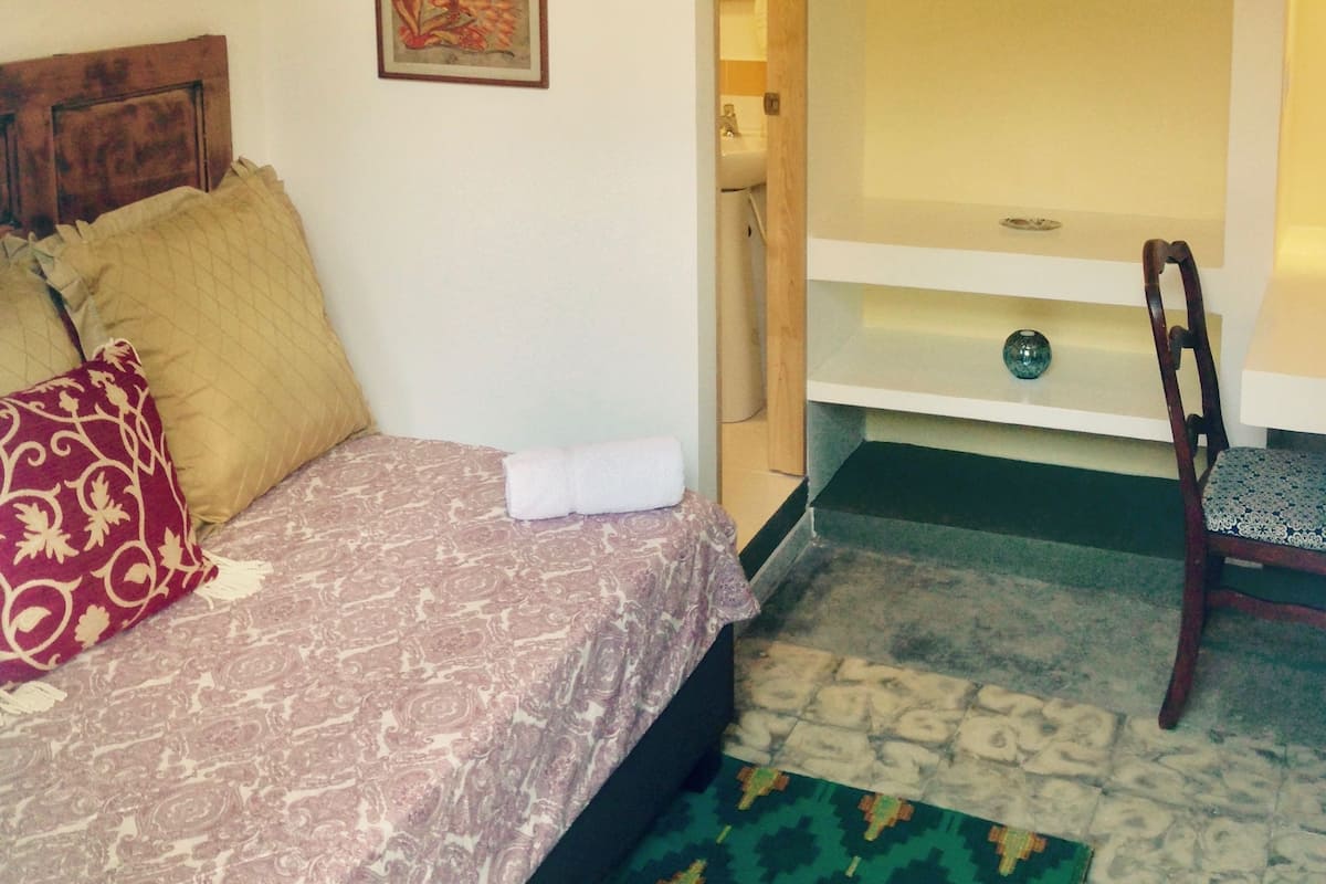 A bedroom with a bed, a ceiling fan, a nightstand, a chair, a potted plant, and a bathroom visible through an open door.