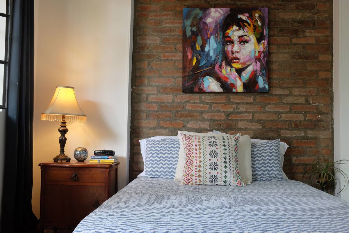 A bedroom with a brick wall, a bed with patterned bedding, a nightstand with a lamp and books, and a colorful painting on the wall.