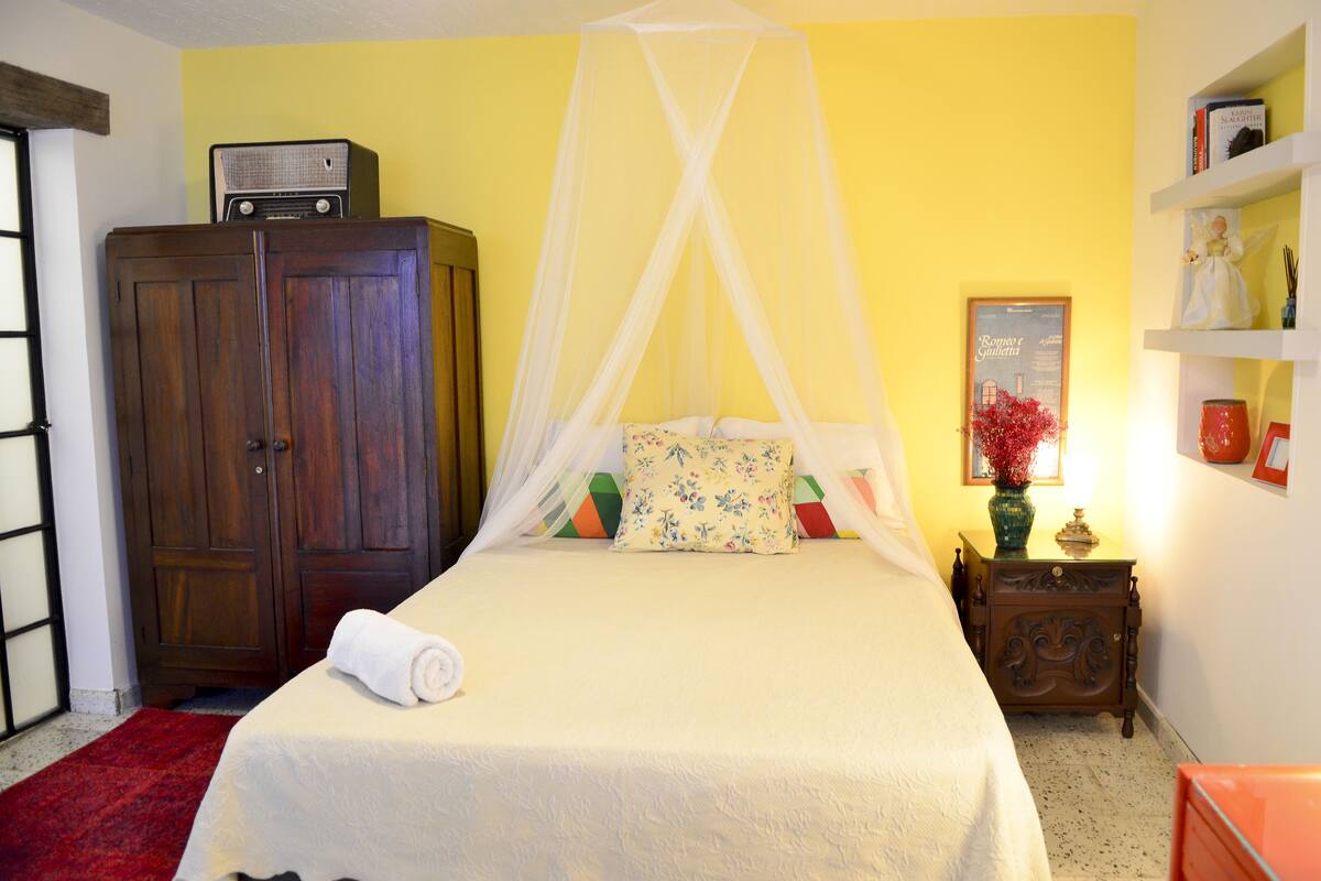 A bedroom with a bed, a wooden wardrobe, a nightstand with a vase, and a wall with a framed picture.