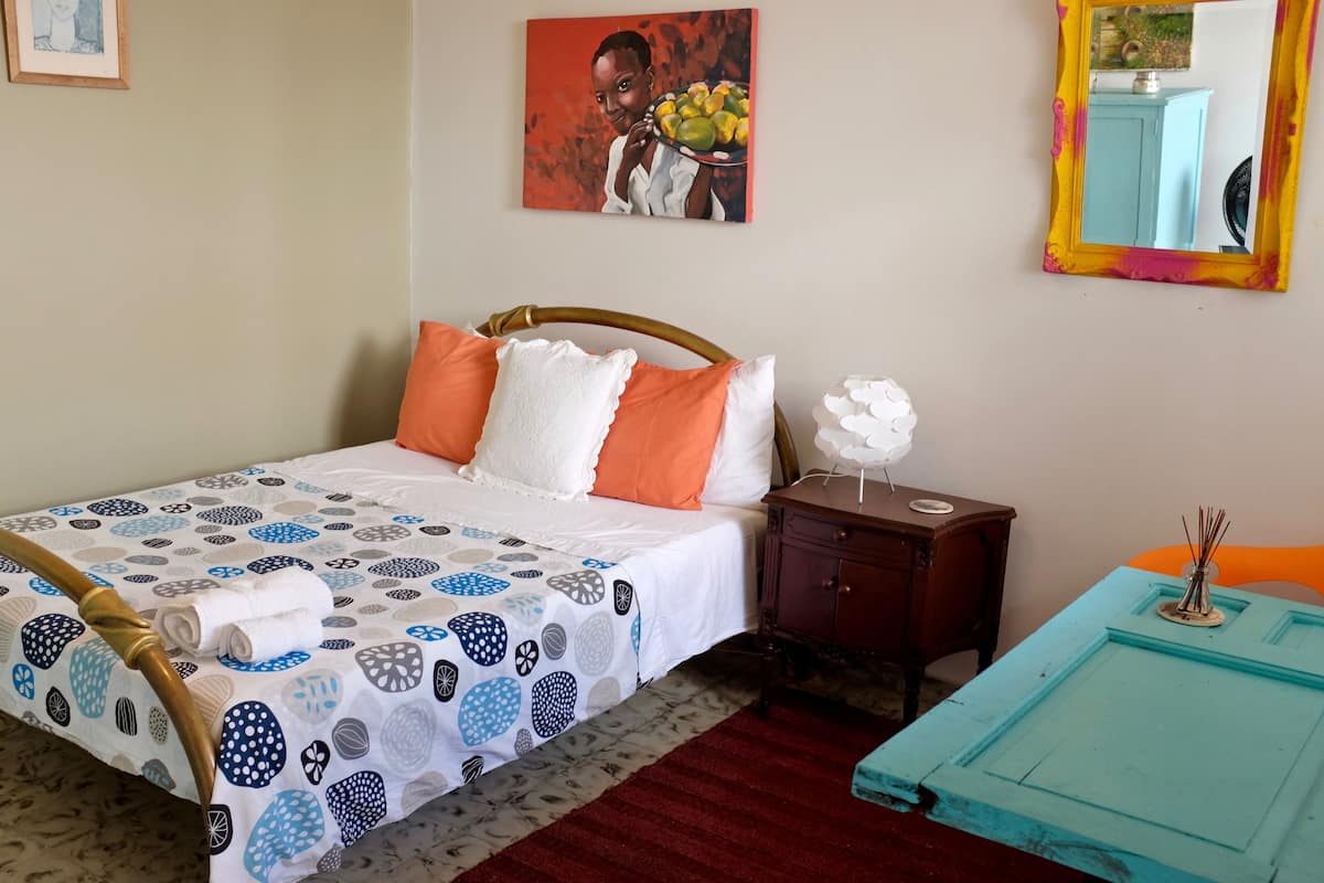 A neatly made bed with patterned bedding, a bedside table with a vase, and a mirror with a colorful frame.