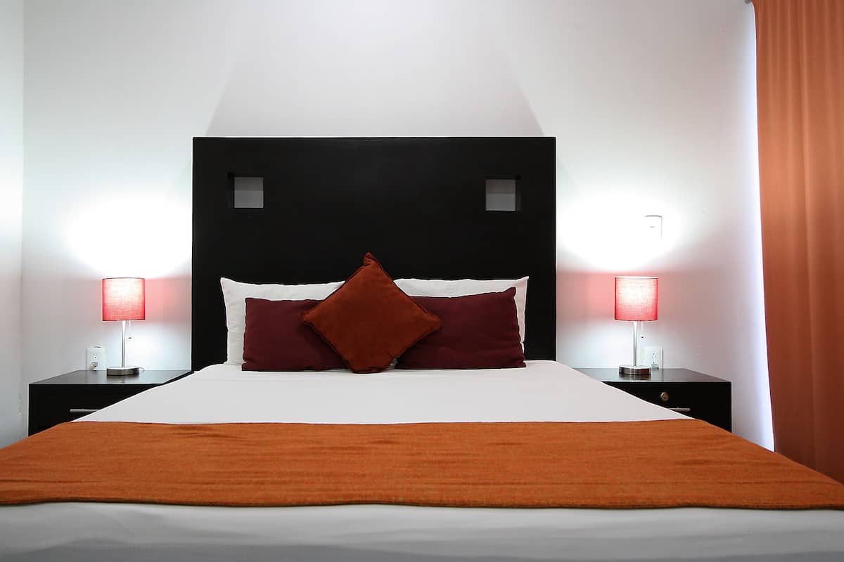 Una habitación de hotel con una cama grande, dos lámparas de noche, una manta roja y un cabecero oscuro.