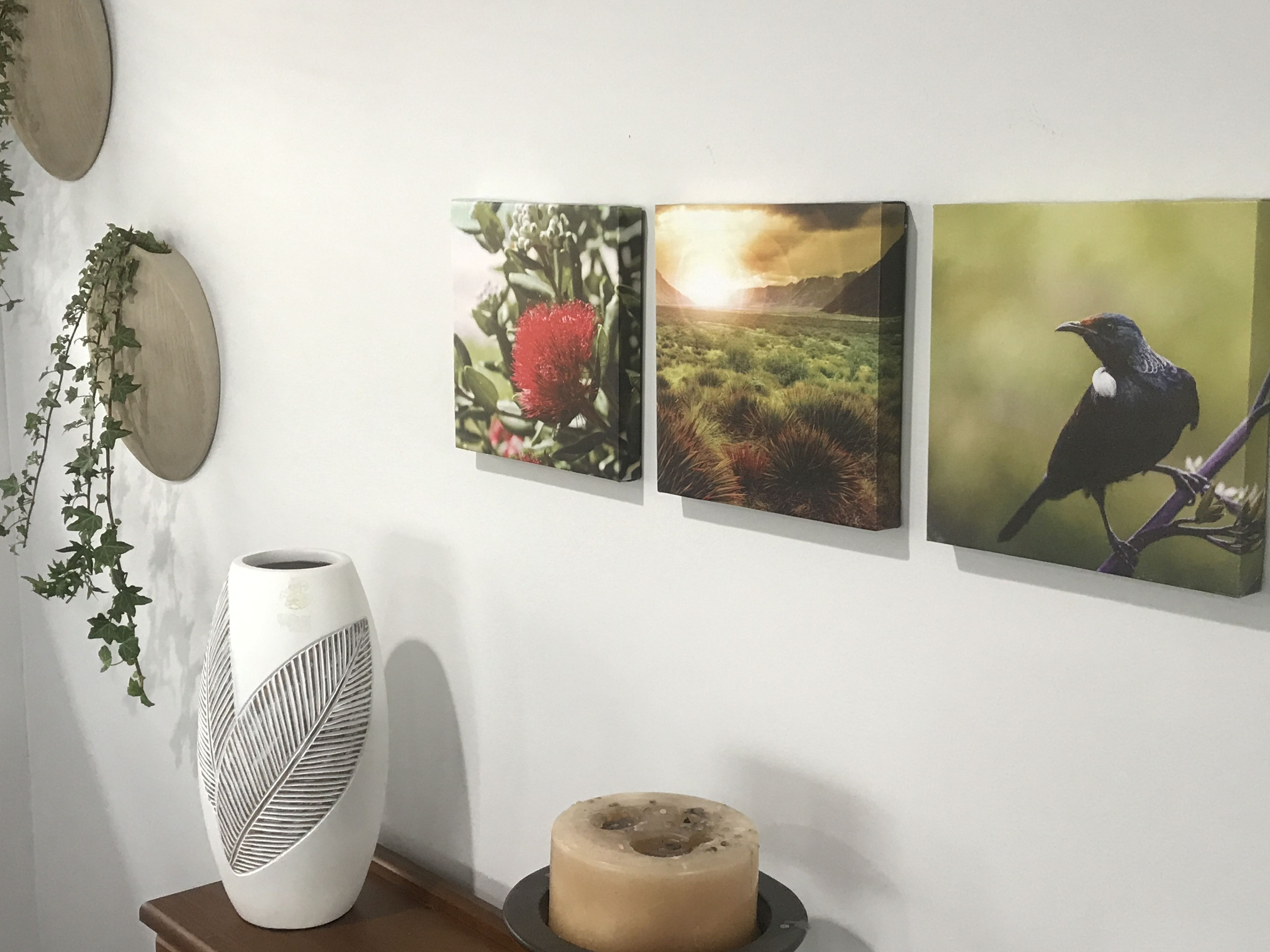 A wall with three framed pictures, a white vase, and a small wooden table with a candle.