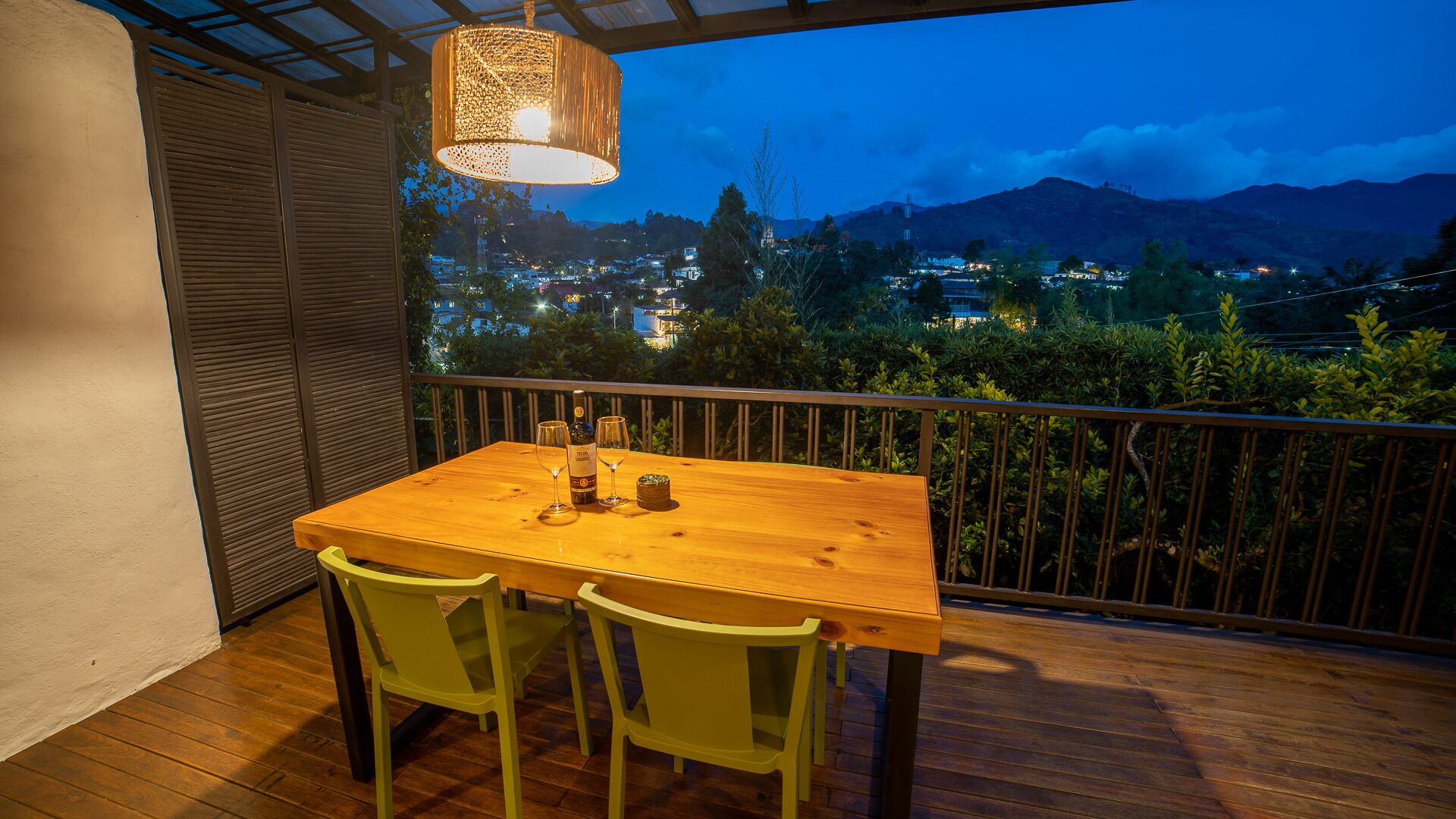 Una mesa de madera con dos sillas en un balcón con vistas a un paisaje urbano al atardecer.
