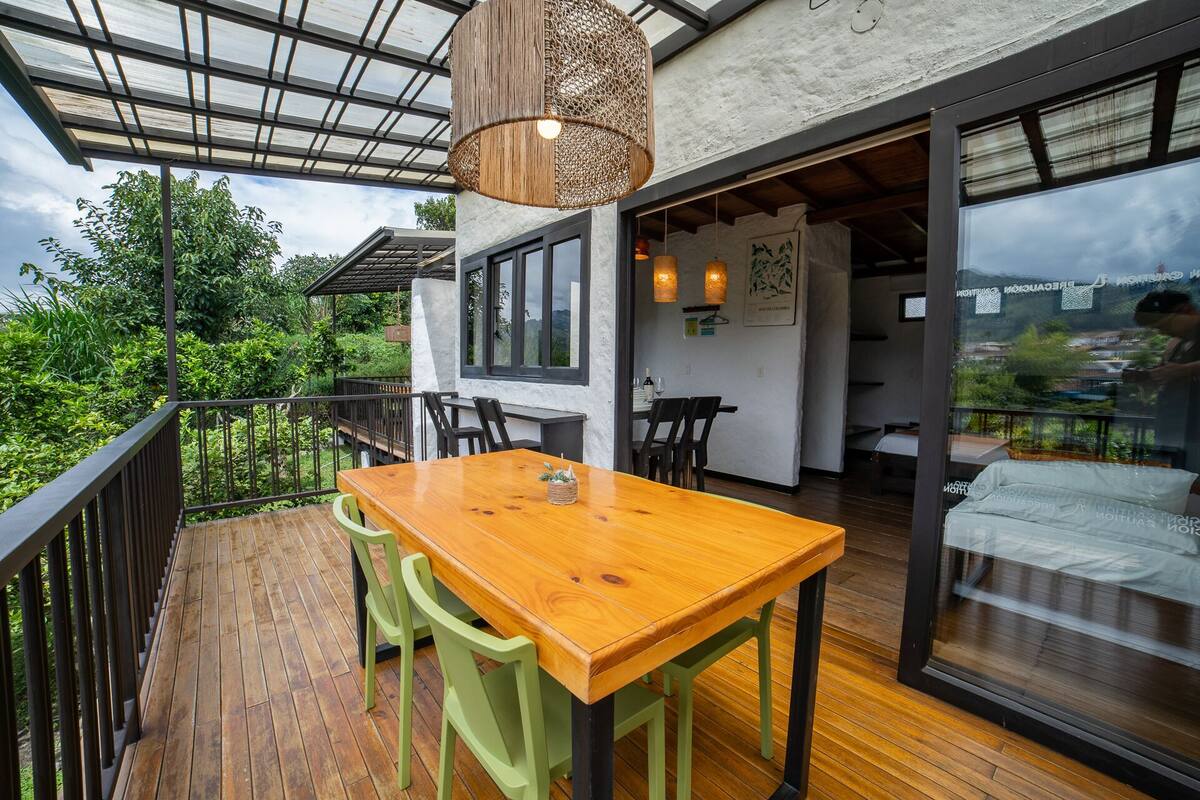 Eine Holzterrasse mit Tisch und Stühlen, einer hängenden Leuchte und Blick auf Grünflächen sowie ein Gebäude im Hintergrund.