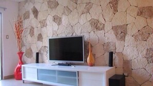 A modern living room with a stone wall, a flat-screen TV on a white cabinet, and a red vase with branches. - Casa Venecia (Playa del Carmen)