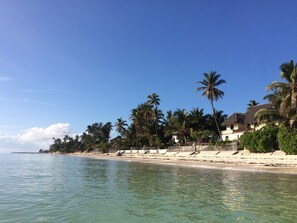 A beach with clear turquoise water, palm trees, and a building with a thatched roof.