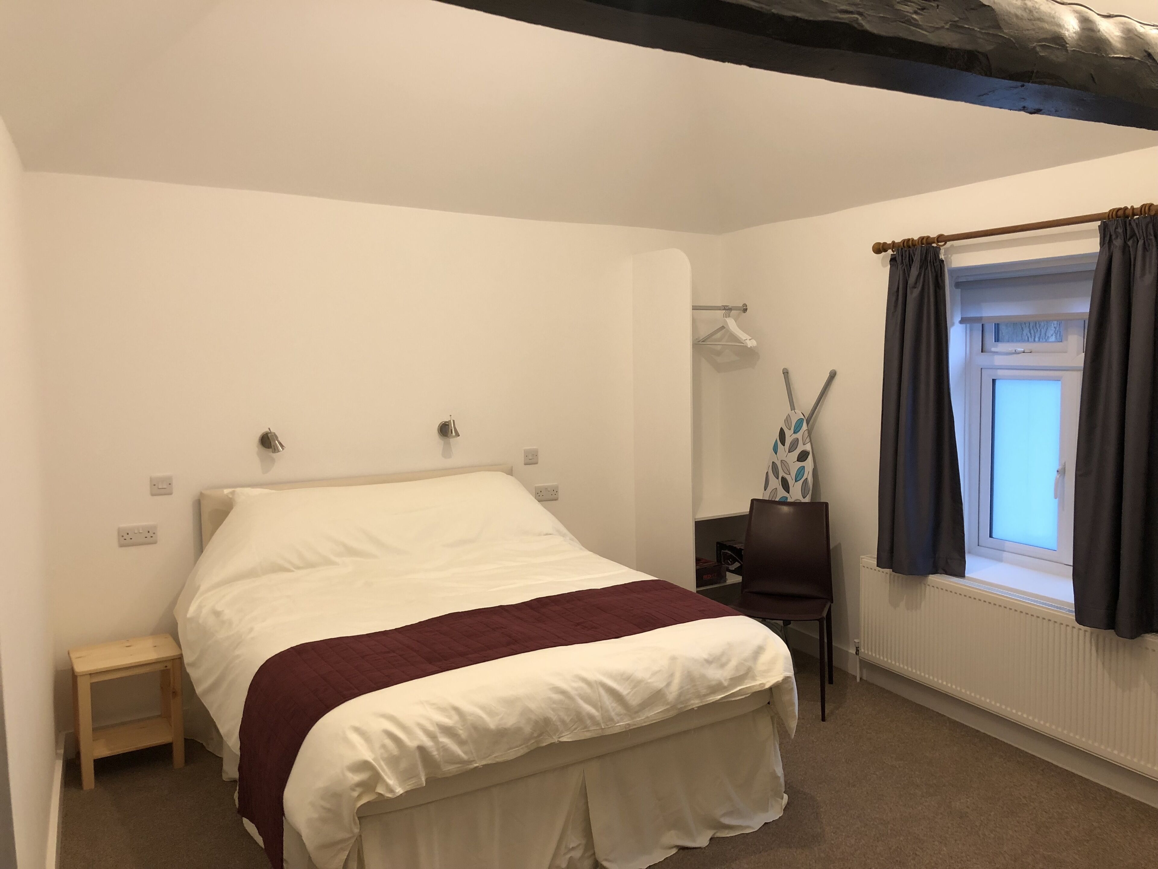A bedroom with a bed, a small bedside table, a chair, a window with curtains, and a radiator.