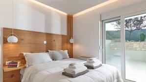 A modern bedroom with a wooden headboard, a large bed with white bedding, two bedside tables with lamps, and a view of the outdoors through a sliding glass door. - Suitable For Families With Children, Groups Of Friends, Professionals, Friends (Rethimno)