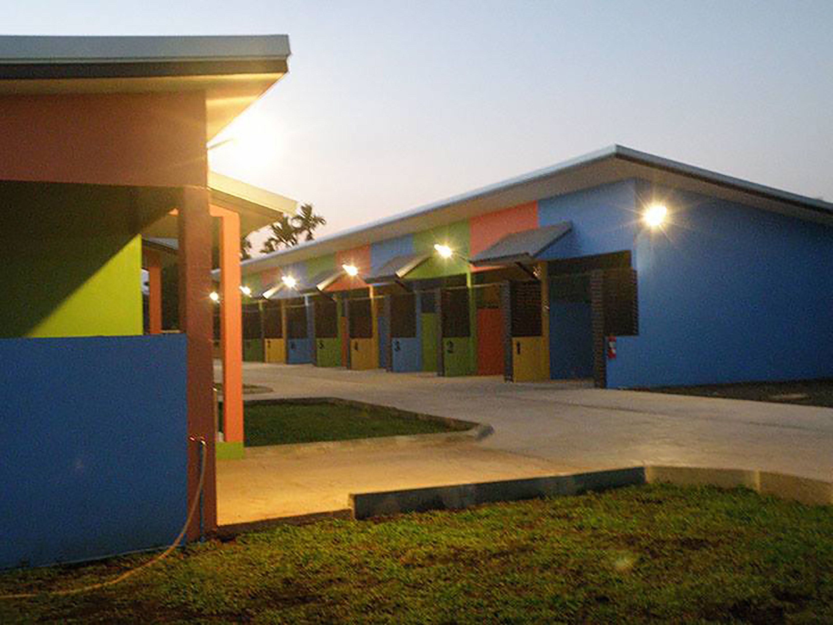 A building with multiple rooms, each with a distinct color. There is a covered outdoor area with a roof and a pathway leading to the entrance.