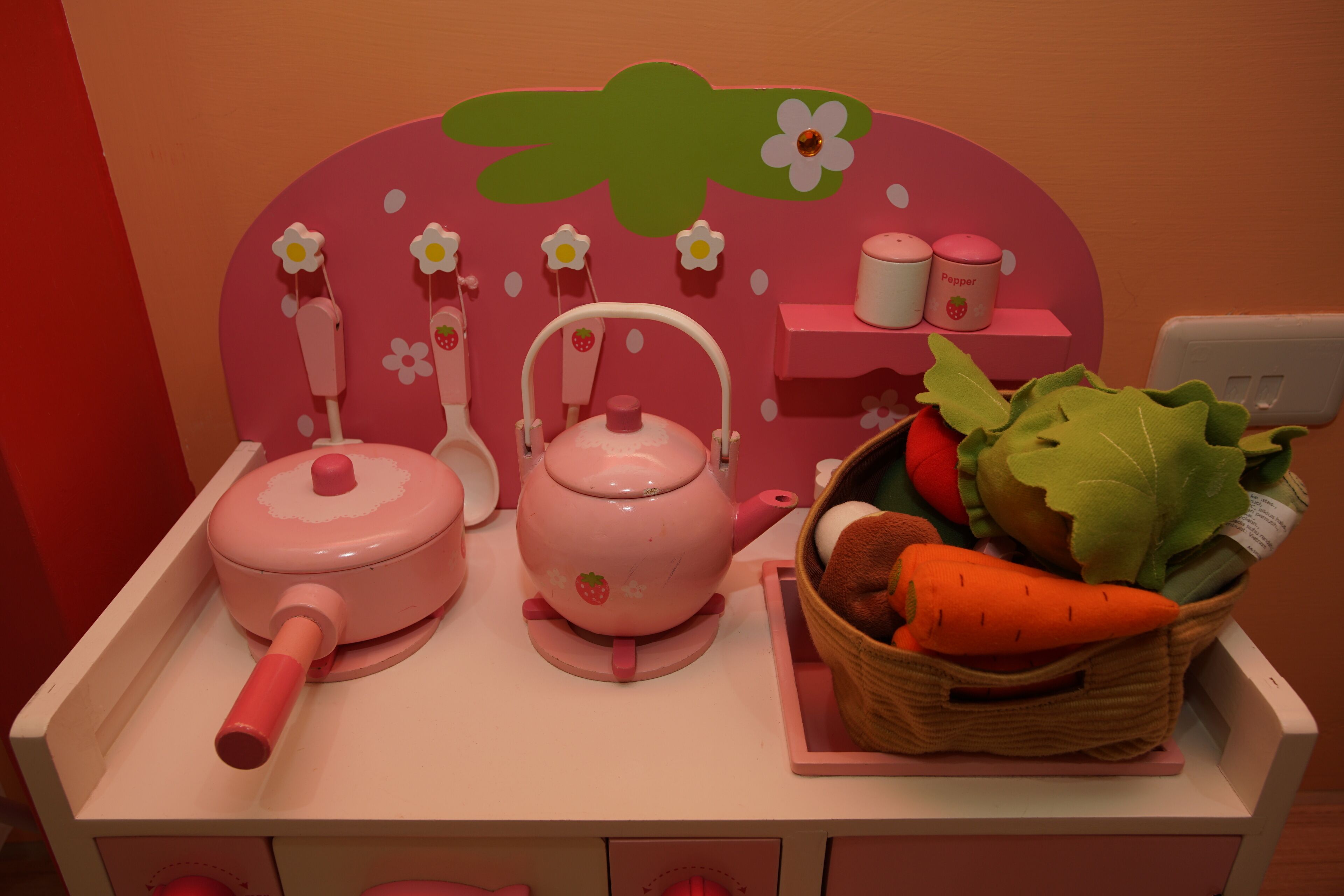 A kitchen setup with a pink theme, including a teapot, a kettle, and a basket of vegetables.