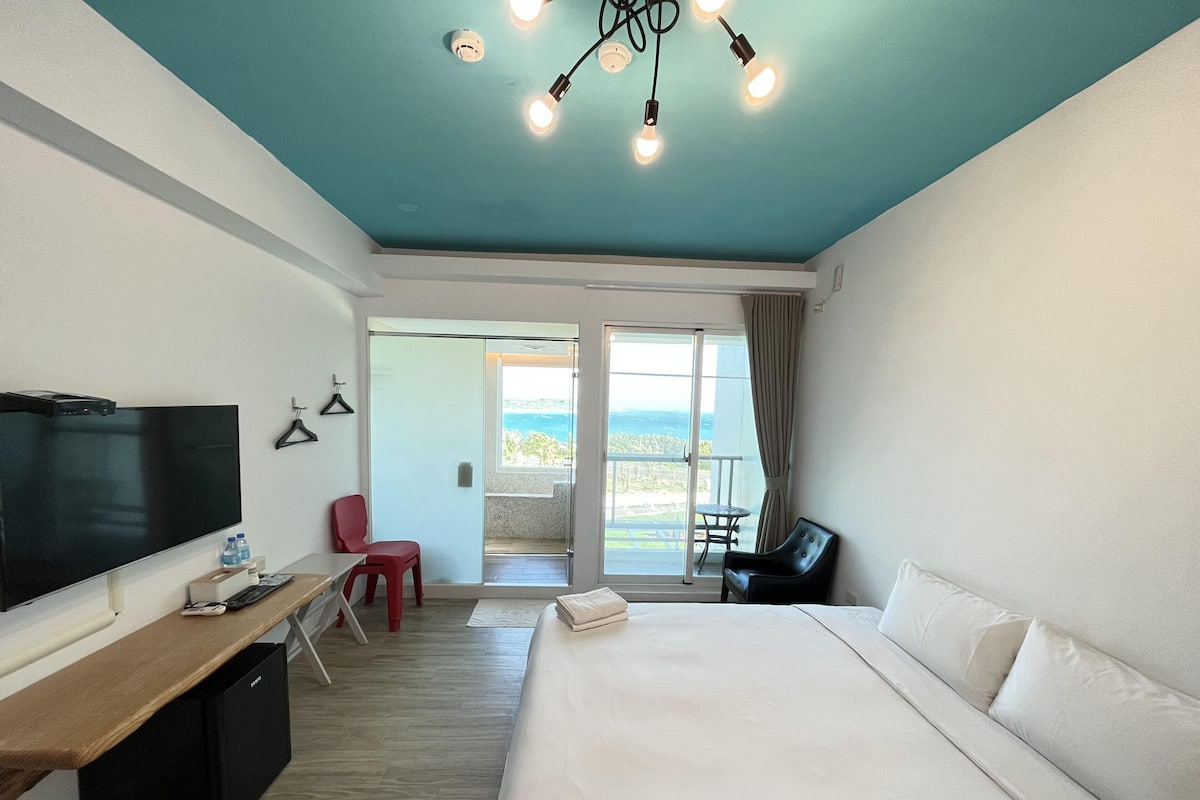A hotel room with a large bed, a desk with a flat-screen TV, a red chair, a black leather chair, and a balcony with a view.