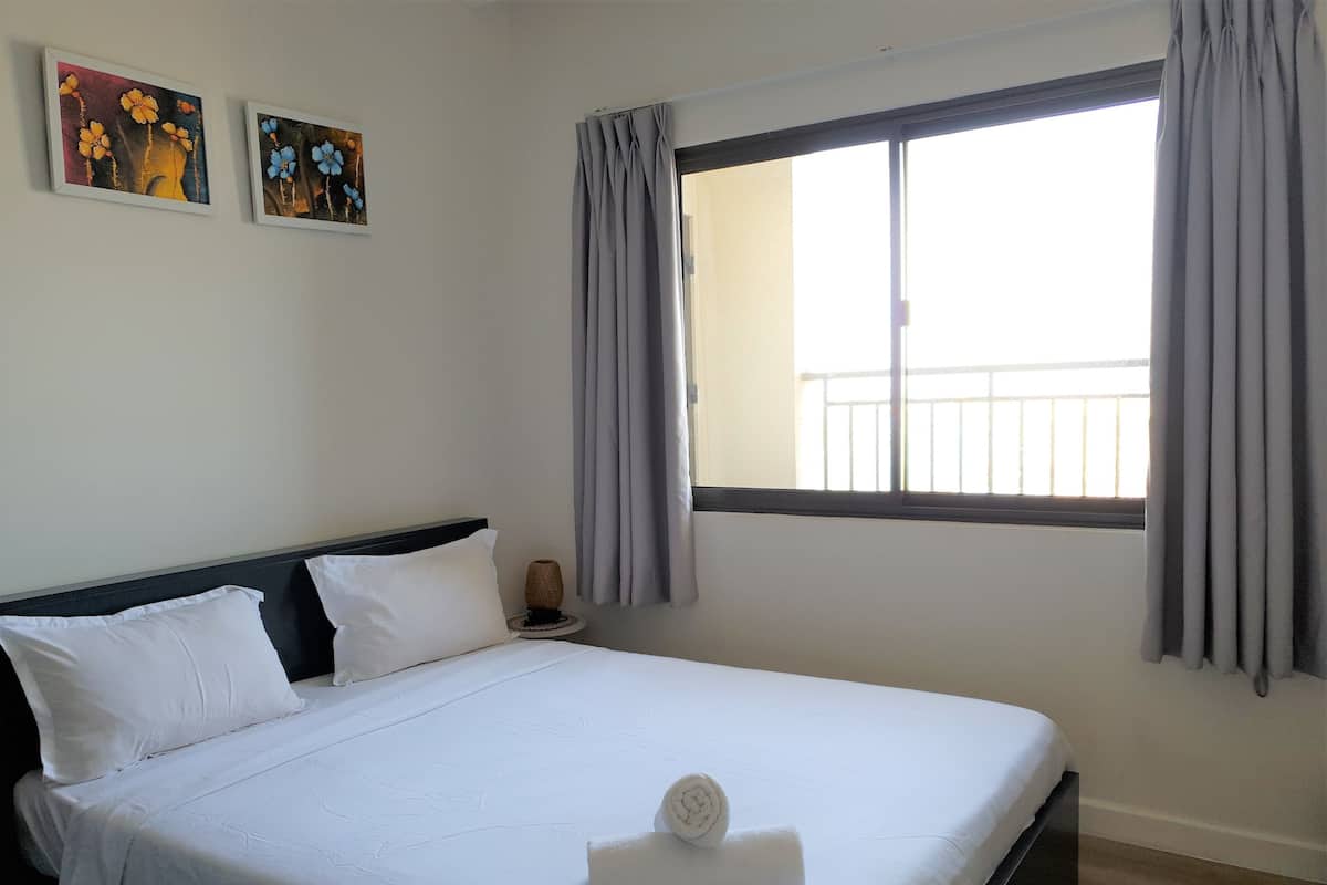 Cama bem arrumada com lençóis brancos e uma toalha enrolada, mesa de cabeceira com abajur, e duas obras de arte emolduradas na parede.