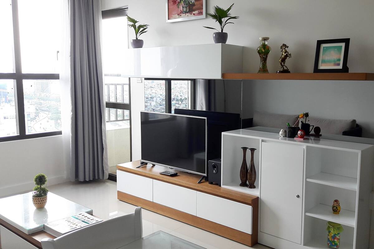 Uma sala de estar moderna com televisão de tela plana, armários brancos, uma pequena mesa de centro e uma janela com vista para a cidade.