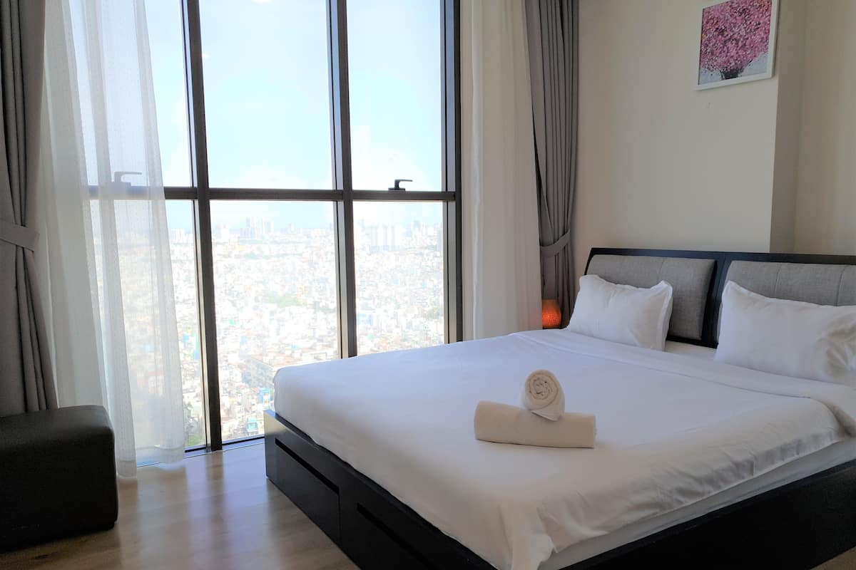 Quarto de hotel com uma cama grande, vista da cidade através de janelas do chão ao teto, uma mesa pequena e um quadro pendurado na parede.