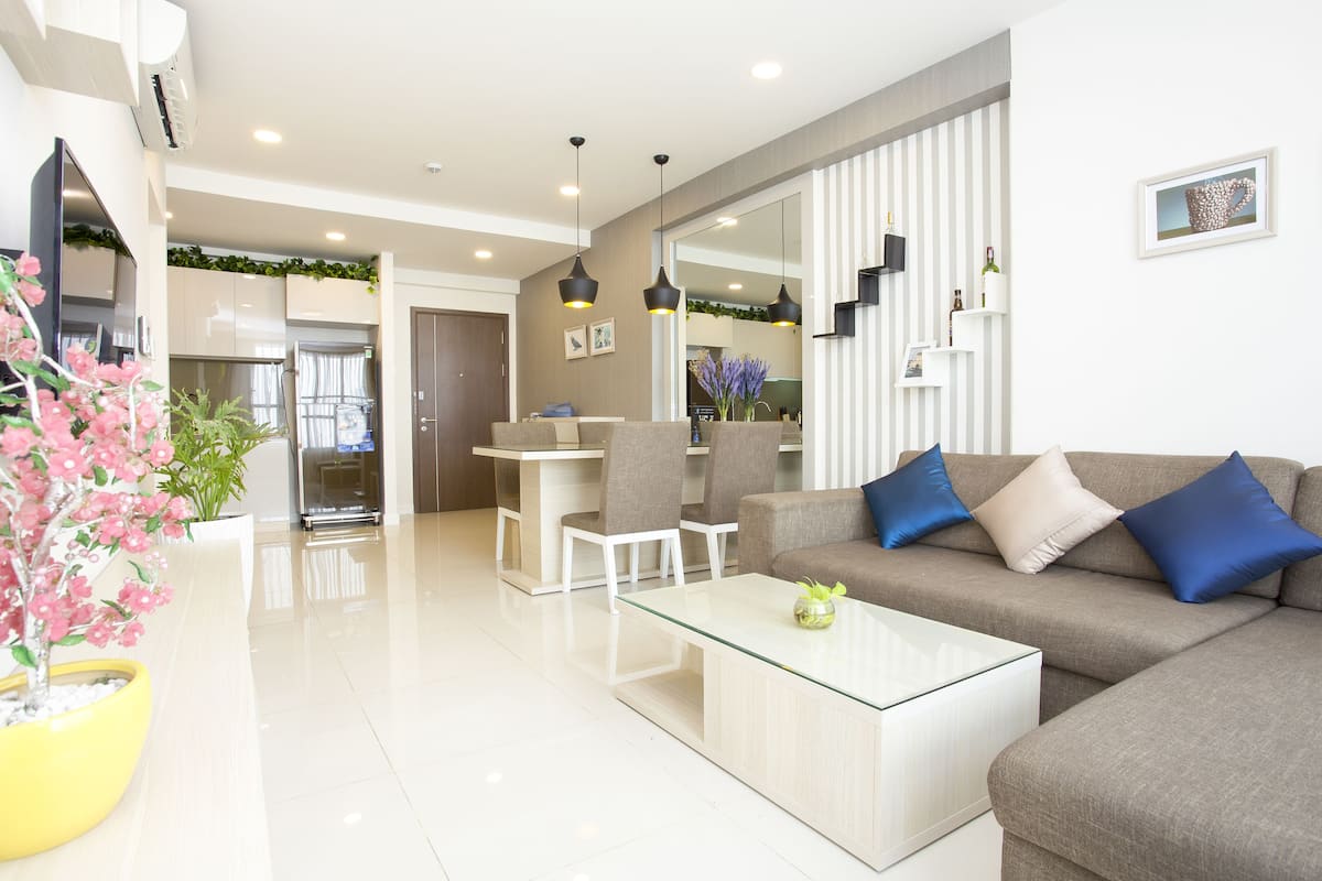 Uma sala de estar moderna com uma mesa de centro de vidro, um sofá cinza com almofadas azuis e uma área de jantar com cadeiras brancas.