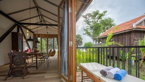 A wooden deck with a bench and a table, a wooden dining area, and a view of a red-roofed building and greenery. - Kampot River Residence (Kampot)