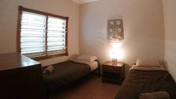 A small, dimly lit room with two single beds, a nightstand with a lamp, and a window with blinds.