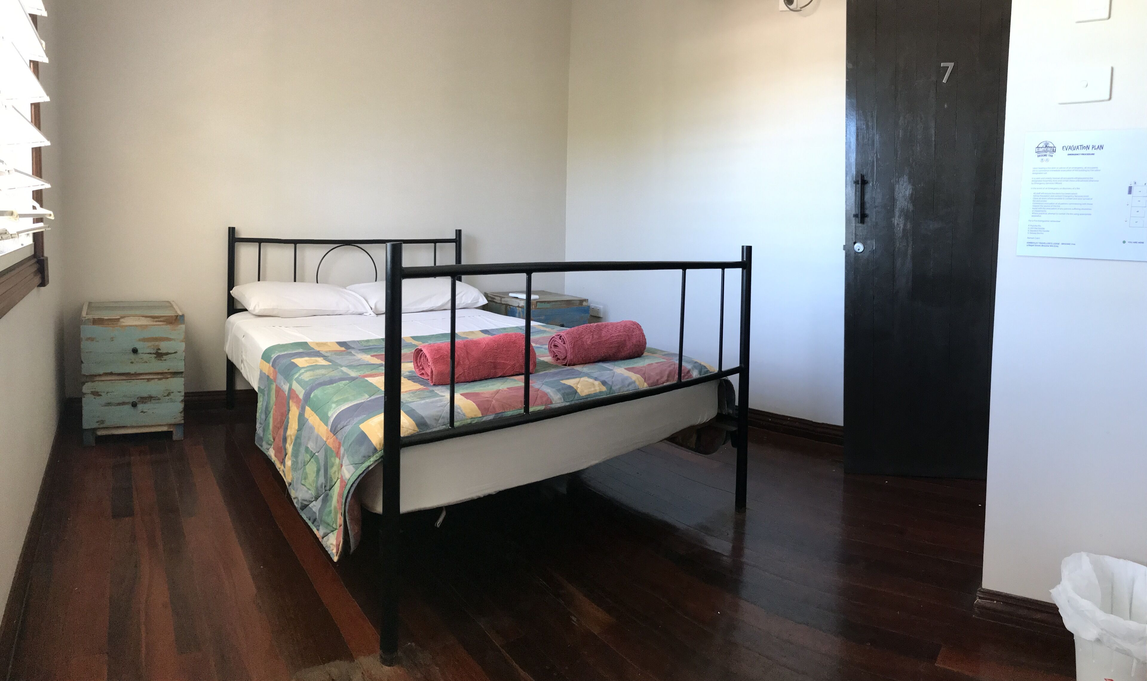 A single bed with a metal frame, a colorful quilt, and a rolled-up towel. There is a small wooden dresser with drawers and a white trash bin.