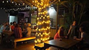 Um deck de madeira com mesas e bancos, rodeado por luzes de corda e uma árvore iluminada.