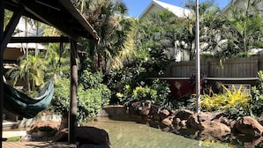Área de piscina com água cristalina, cercada por pedras e palmeiras, e uma rede pendurada em uma estrutura.