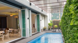 A modern outdoor pool area with a wooden deck, glass doors leading to a seating area, and a green wall with plants.