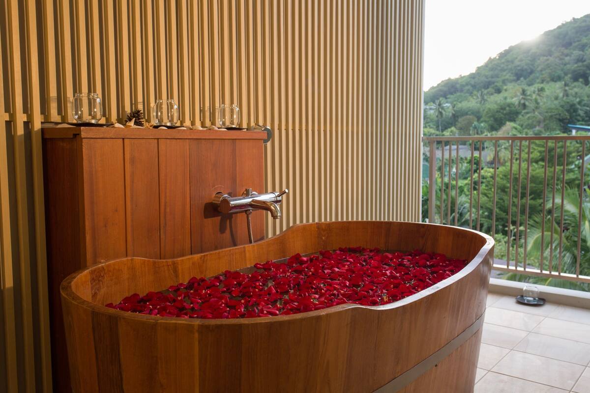 Una bañera de madera llena de pétalos de rosa roja, un armario de madera con cristalería, y un balcón con vistas a la vegetación.