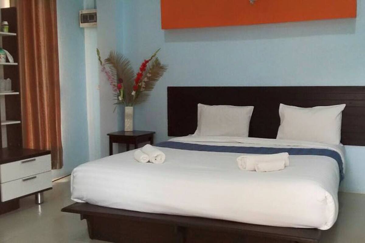 Una cama bien hecha con sábanas blancas y dos toallas enrolladas, una mesita de noche con un florero, y un cabecero oscuro frente a una pared azul claro.