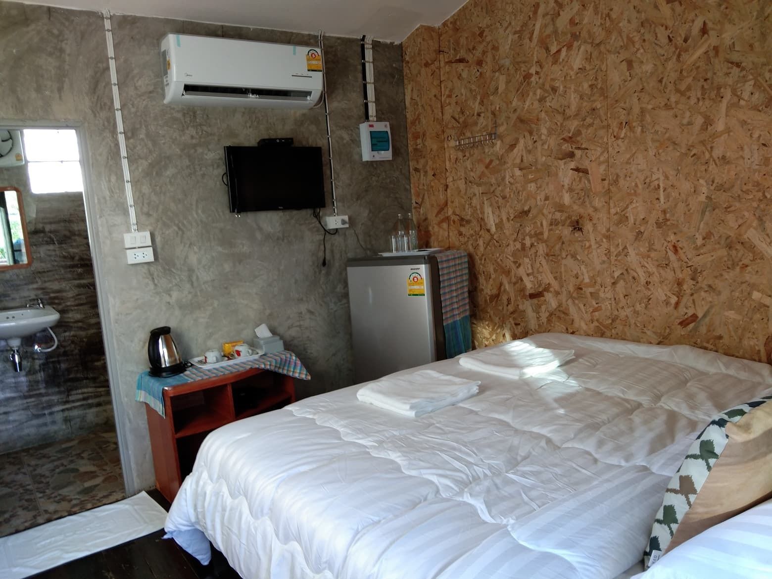 Una habitación con cama, una mesita con hervidor, refrigerador y televisor montado.