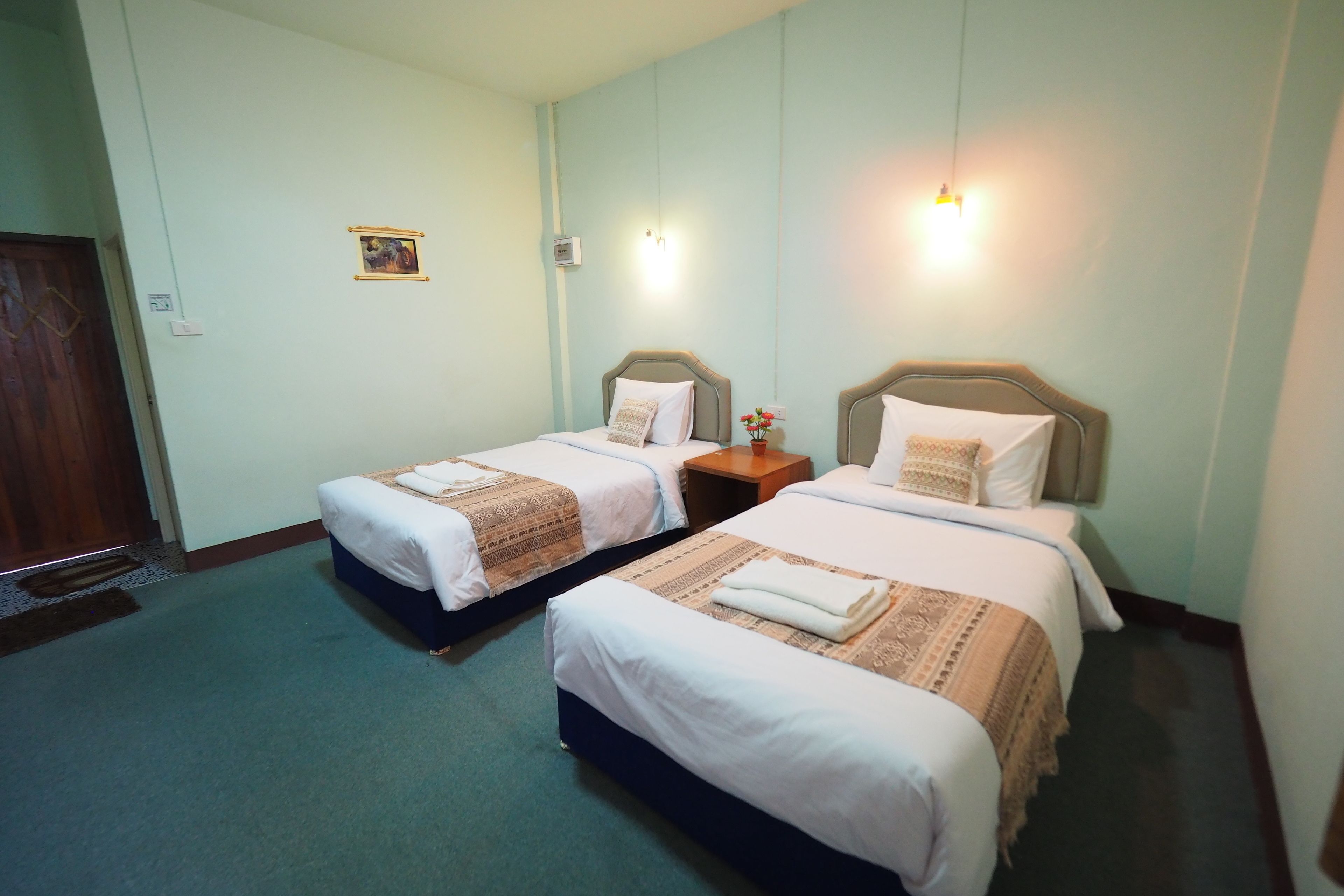 A hotel room with two beds, a wooden door, a small bedside table with a lamp, and a wall-mounted light fixture.