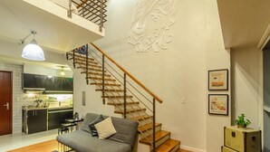 A modern living room with a grey sofa, a wooden staircase, a kitchen in the background, and a decorative wall piece. - Eton Residences by StayHome Asia (Makati)