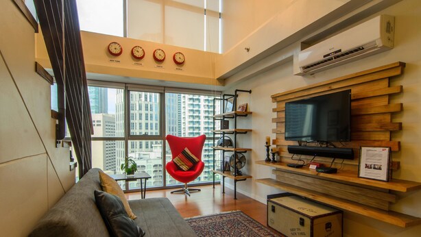 A modern living room with a large TV, a red chair, a sofa, and a city view through large windows. - Eton Residences by StayHome Asia (Makati)