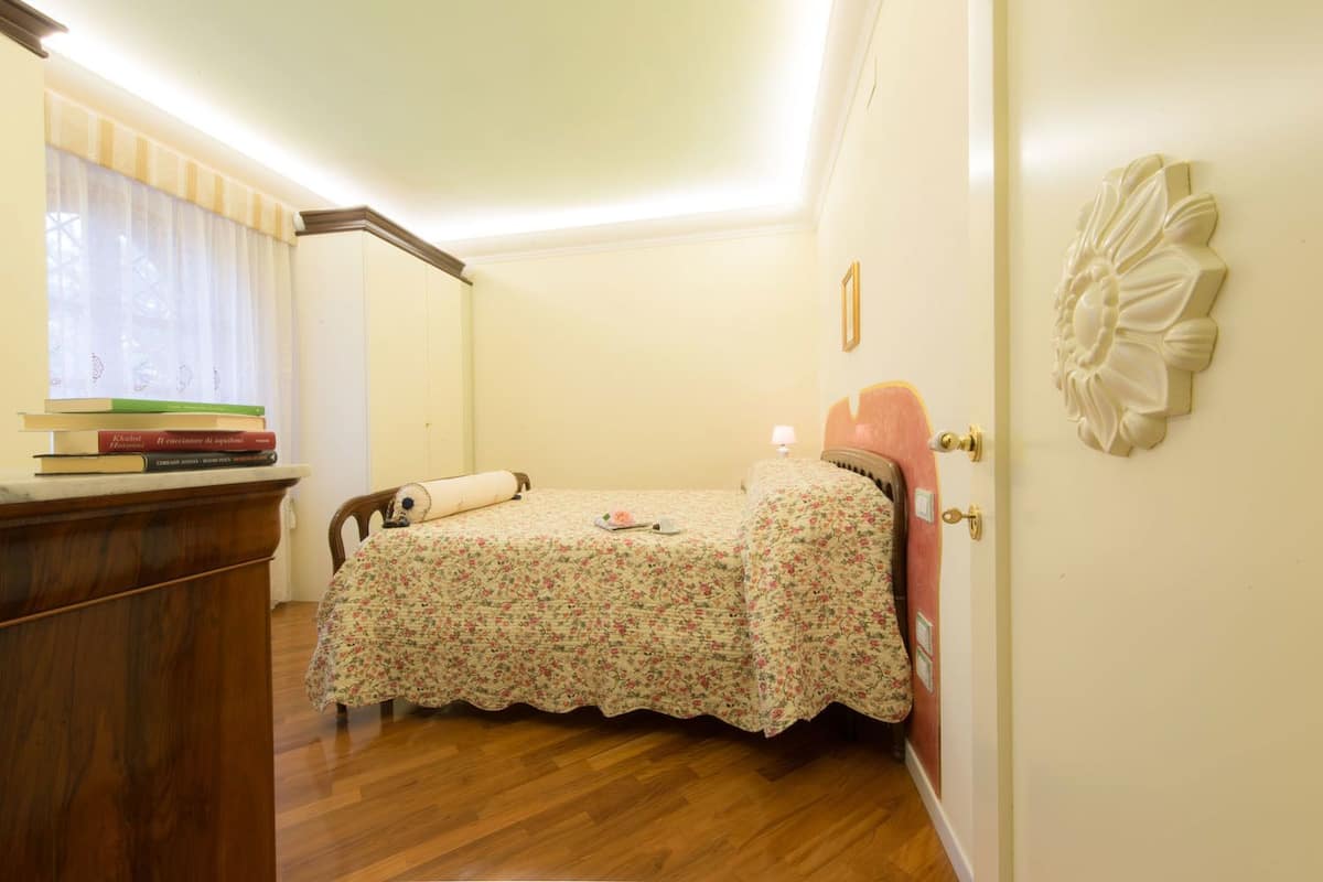 Une petite chambre à coucher accueillante, avec un plancher en bois, un lit recouvert d’une literie à motifs floraux, une table de chevet avec des livres et une fenêtre agrémentée de rideaux.